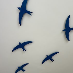 May include: A collection of six blue bird-shaped wall decorations. The birds are in various positions, with wings spread, as if in flight. The background is a plain white surface, creating a contrast with the dark blue birds.