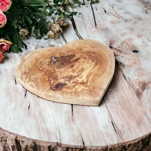 Peut inclure: Une planche à découper en bois en forme de cœur, en bois d'olivier, avec un motif de grain de bois naturel. La planche est posée sur une surface en bois rustique.