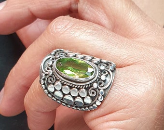 Pode incluir: Um anel de prata com uma gema verde ovalada engastada em um design detalhado. O anel é usado em um dedo.
