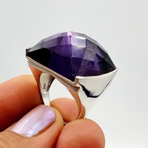 Könnte beinhalten: Ein Ring aus Sterlingsilber mit einem großen, facettierten, tiefvioletten Amethyst-Edelstein. Der Stein ist in einer geometrischen, vielflächigen Form geschliffen und in eine polierte Silberschiene gefasst, die innen einen 925er Stempel zeigt.