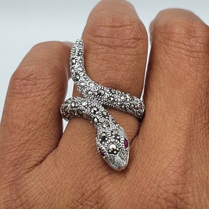 Peut inclure: Bague serpent en argent ornée de détails en marcassite et d'un œil en pierre rouge. La bague est enroulée autour d'un doigt, mettant en valeur son design enroulé. La tête du serpent est ornée de petites pierres rondes.
