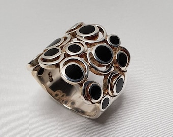 May include: A silver ring with a wide band featuring multiple black circles in a geometric pattern.