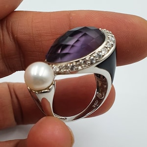 Könnte beinhalten: Ein silberner Ring mit einem großen violetten Edelstein, der von kleineren weißen Edelsteinen umgeben ist. Der Ring hat auch eine weiße Perle an der Seite.