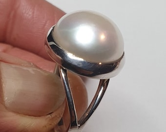 Może przedstawiać: Srebrny pierścionek z dużym, białym perłą osadzonym w oprawie typu bezel. Pierścionek ma prosty, elegancki design.