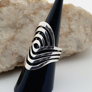 May include: A silver ring with a geometric design. The ring is on a black ring holder and a light brown rock.