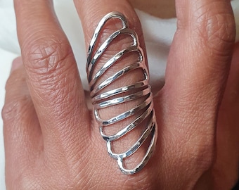 May include: A silver-colored ring with a unique design. The ring features a series of curved, parallel bars that create an elongated, fan-like shape. The metal has a textured, hammered appearance. The ring is worn on a finger.