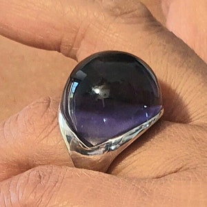 Könnte beinhalten: Ein auffälliger Ring mit einem großen, kuppelförmigen Edelstein in dunklen Lilatönen, gefasst in einem silbernen Band. Der Edelstein hat einen Farbverlauf. Das Design des Rings ist modern und auffällig.