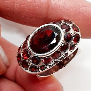 Può includere: Un anello d'argento con una grande pietra ovale rossa circondata da pietre rosse più piccole.