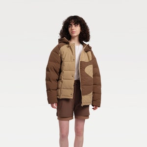 May include: A brown and tan hooded puffer jacket with a distinctive wave pattern. The model is wearing a white top, brown shorts, white socks, and trainers. The jacket features a quilted design.