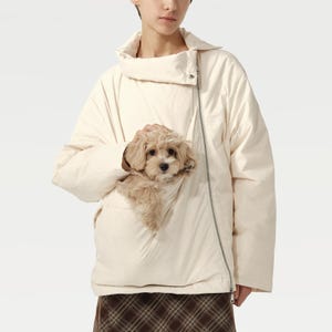 Puede incluir: Chaqueta acolchada color crema con cremallera diagonal y cuello alto. La chaqueta tiene un gran bolsillo delantero que sostiene un perro pequeño, esponjoso y marrón claro. El perro mira directamente a la cámara.