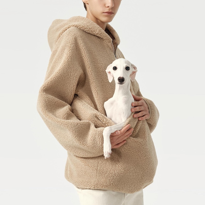Pu&ograve; includere: Una felpa con cappuccio in pile sherpa color cammello con una tasca a marsupio, che tiene in braccio un piccolo cane bianco. Il cane ha grandi orecchie dritte e guarda la fotocamera. La felpa ha una chiusura a mezza zip.