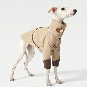 May include: A tan dog coat with brown trim and accents. The coat features a high collar, button closures, and brown cuffs. The dog is a light color, and the coat is tailored to fit. The dog is standing on a white background.