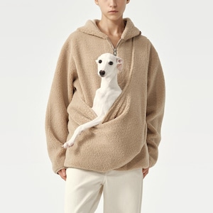 Könnte beinhalten: Beigefarbener Sherpa-Fleece-Hoodie mit halbem Reißverschluss. Ein kleiner weißer Hund liegt in der Vordertasche des Hoodies. Der Hund hat einen langen Hals und spitze Ohren. Die Person trägt eine weiße Hose.