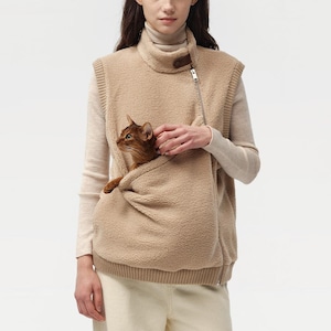 Puede incluir: Un chaleco de forro polar beige con cuello alto y cremallera diagonal. El chaleco tiene un gran bolsillo delantero que contiene un gato marrón. El gato mira hacia la izquierda. El chaleco está hecho de un material suave y texturizado.