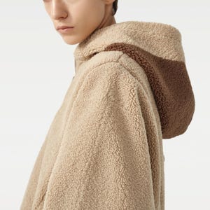 Pu&ograve; includere: Giacca con cappuccio in pile beige con un accento marrone sul cappuccio. La giacca ha un aspetto morbido e strutturato. L'immagine &egrave; un primo piano della giacca e del cappuccio.