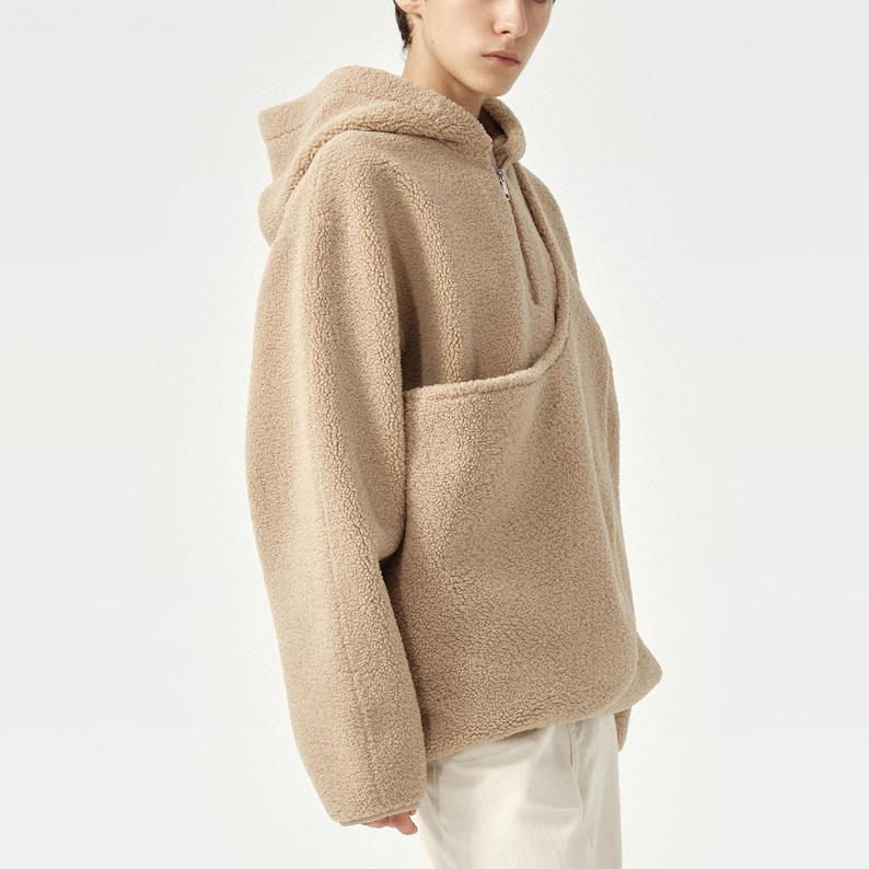 Pu&ograve; includere: Una felpa con cappuccio beige con mezza zip e un'ampia tasca frontale. La felpa &egrave; realizzata in un materiale morbido e strutturato. Il modello indossa pantaloni bianchi. Il cappuccio &egrave; visibile.