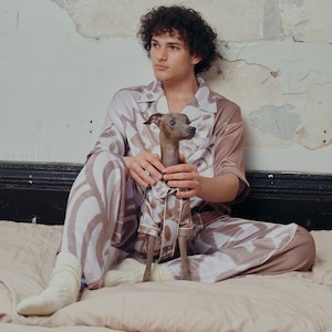 Op de afbeelding: Een persoon in een bruin en wit gepatroneerd pyjama-set zit op een bed met een kleine grijs-bruine hond. De persoon houdt de hond op schoot.