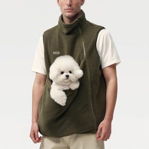 Puede incluir: Un chaleco de forro polar verde oliva con cuello alto, cierre de cremallera asimétrico y un bolsillo frontal grande. Un pequeño perro Bichon Frise blanco está colocado dentro del bolsillo frontal, con su cabeza y patas delanteras visibles.