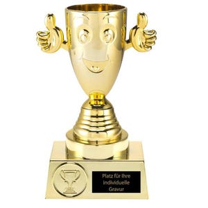 May include: A gold-colored trophy with a smiling face and thumbs-up hands. The trophy sits on a square base with a small plaque that reads "Platz für Ihre individuelle Gravur". The trophy is designed to be personalized.