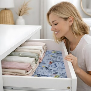 May include: A white chest of drawers with an open drawer containing folded pastel linens. A blue patterned drawer liner with toucans and tropical leaves is visible. A person is looking into the drawer.