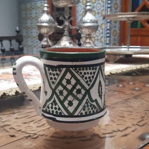 May include: A white ceramic mug with a green and black geometric pattern. The mug has a white handle and a green rim. The mug is sitting on a glass table. The mug is likely used for drinking beverages.