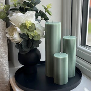 a black tray with three green candles on it, placed on a windowsill. The tray also holds a vase filled with white flowers and greenery.