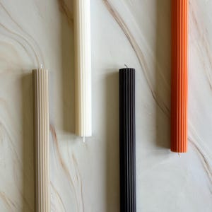 four candles of different colors - white, black, orange, and beige - arranged on a marble surface.