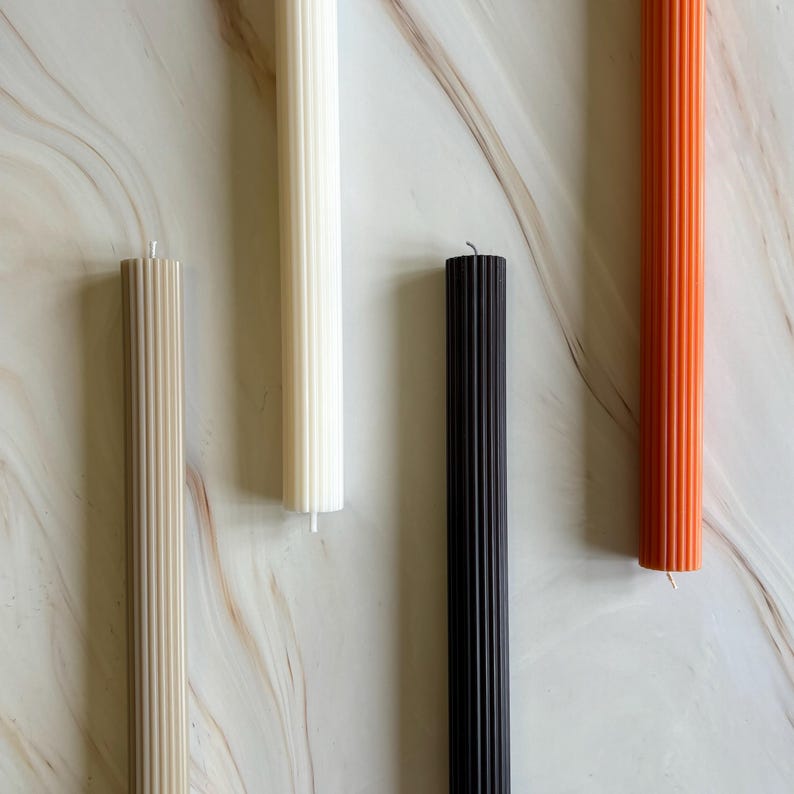 four candles of different colors - white, black, orange, and beige - arranged on a marble surface.