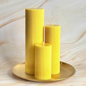 Three tall, cylindrical yellow candles with a textured appearance are displayed on a gold plate against a marble background.