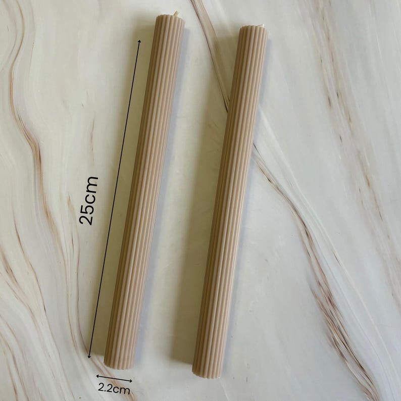 two cylindrical objects, possibly candles, placed on a marble surface. The objects appear to be made of a light-colored material, possibly wax or paraffin. The image also includes measurements, indicating that the objects are approximately 25 cm in length.
