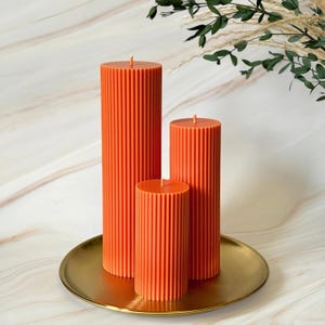 Three tall orange candles with a textured surface are displayed on a gold plate, with a plant branch visible in the background.