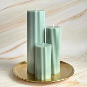 Three tall, cylindrical candles in a light green color are displayed on a gold plate against a marble background.