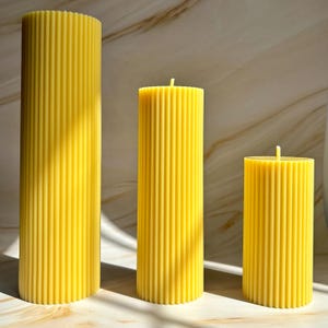 Three tall, cylindrical candles with a yellow color are placed on a marble surface, casting shadows on the marble.