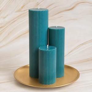 Three tall, cylindrical candles in a teal color are displayed on a gold plate against a marble background.