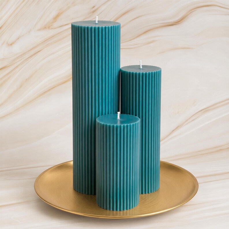 Three tall, cylindrical candles in a teal color are displayed on a gold plate against a marble background.
