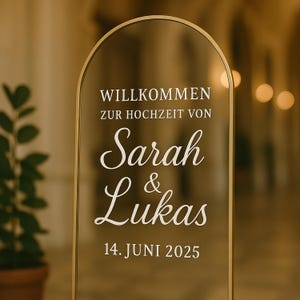 Può includere: Un cartello di benvenuto per matrimoni in acrilico trasparente con una cornice dorata. Il cartello presenta le parole "Willkommen zur Hochzeit von Sarah & Lukas 14. Juni 2025" in caratteri bianchi. Il cartello ha una parte superiore ad arco.