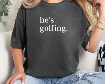 May include: A dark gray oversized t-shirt with the text "he's golfing." printed in white.