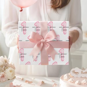 Może przedstawiać: Prezent owinięty r&oacute;żową wstążką, z powtarzającym się wzorem liczby 1 i słowami "Happy Birthday Emma". R&oacute;żowy balon unosi się nad nim. W tle tort urodzinowy i kwiaty.