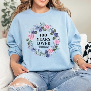 May include: Light blue sweatshirt with a floral wreath design and the text "100 Years Loved Patricia".