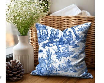 May include: A white pillow with a blue and white toile pattern featuring a pastoral scene with figures and trees. The pillow is sitting on a wicker basket.