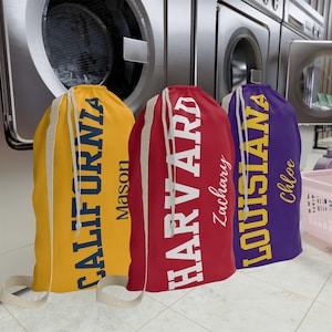 Puede incluir: Tres bolsas de lavandería personalizadas en una lavandería. Las bolsas son amarillas, rojas y moradas, cada una con un nombre de universidad diferente impreso verticalmente en letras blancas en negrita. Cada bolsa también tiene un nombre impreso debajo.