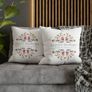May include: Two white throw pillows with floral designs and the text "Bailey Dupont - Wedding Organizer" on a gray couch.
