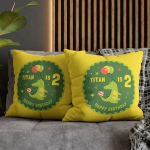 Pode incluir: Duas almofadas amarelas com c&iacute;rculos verdes. Os c&iacute;rculos t&ecirc;m um jacar&eacute; verde segurando bal&otilde;es e um presente. O texto nas almofadas diz "TITAN IS 2 HAPPY BIRTHDAY".