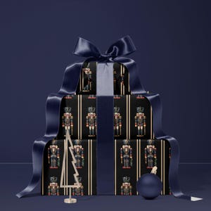 May include: A stack of wrapped gifts with a navy blue ribbon and bow. The wrapping paper features a repeating pattern of nutcracker soldiers on a black background with gold stripes. A small gold Christmas tree and a blue ornament are also present.