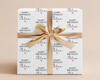 Może przedstawiać: Prezent zapakowany w biały papier z napisem "HAPPY BIRTHDAY Chloe" czarnym pismem, z różowymi serduszkami. Prezent jest zawiązany złotą wstążką i kokardą. Temat: uroczystość.