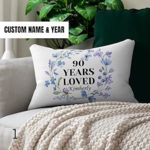 May include: White throw pillow with a floral wreath design and the text "90 Years Loved Kimberly".