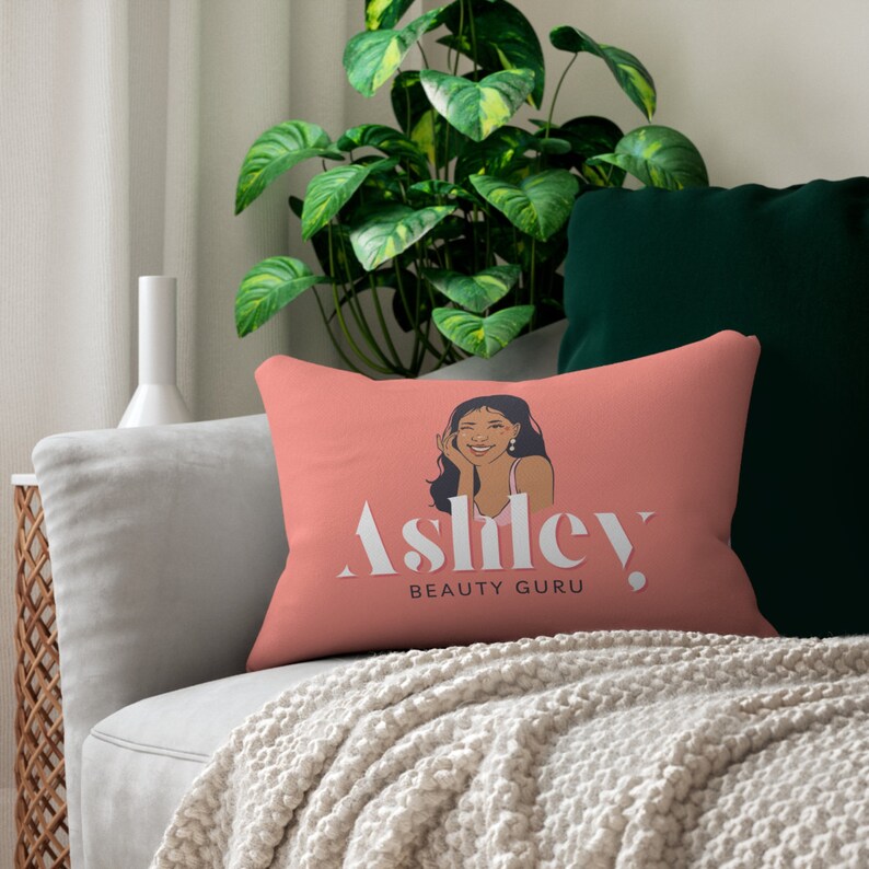 May include: A pink throw pillow with a graphic of a woman with long black hair and the text "Ashley BEAUTY GURU".