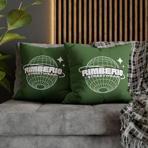 May include: Two green throw pillows with white text that reads "Rimberio Streetwear" and a graphic of a globe with a ring around it. The pillows are on a gray couch with a black and white plaid throw blanket.