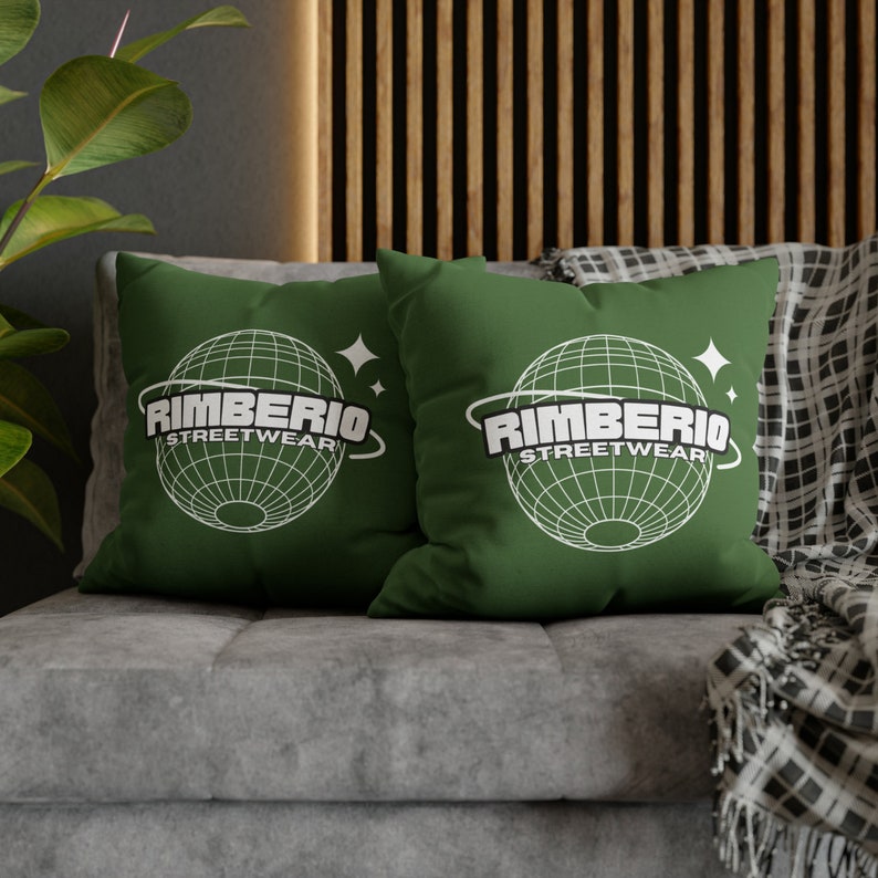 May include: Two green throw pillows with white text that reads "Rimberio Streetwear" and a graphic of a globe with a ring around it. The pillows are on a gray couch with a black and white plaid throw blanket.