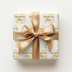Może przedstawiać: Prezent urodzinowy owinięty w biały papier, z powtarzającym się wzorem liczby 10 w kolorze ż&oacute;łtym, słowami "Happy Birthday" i imieniem "Jason". Jest zawiązany złotą wstążką.